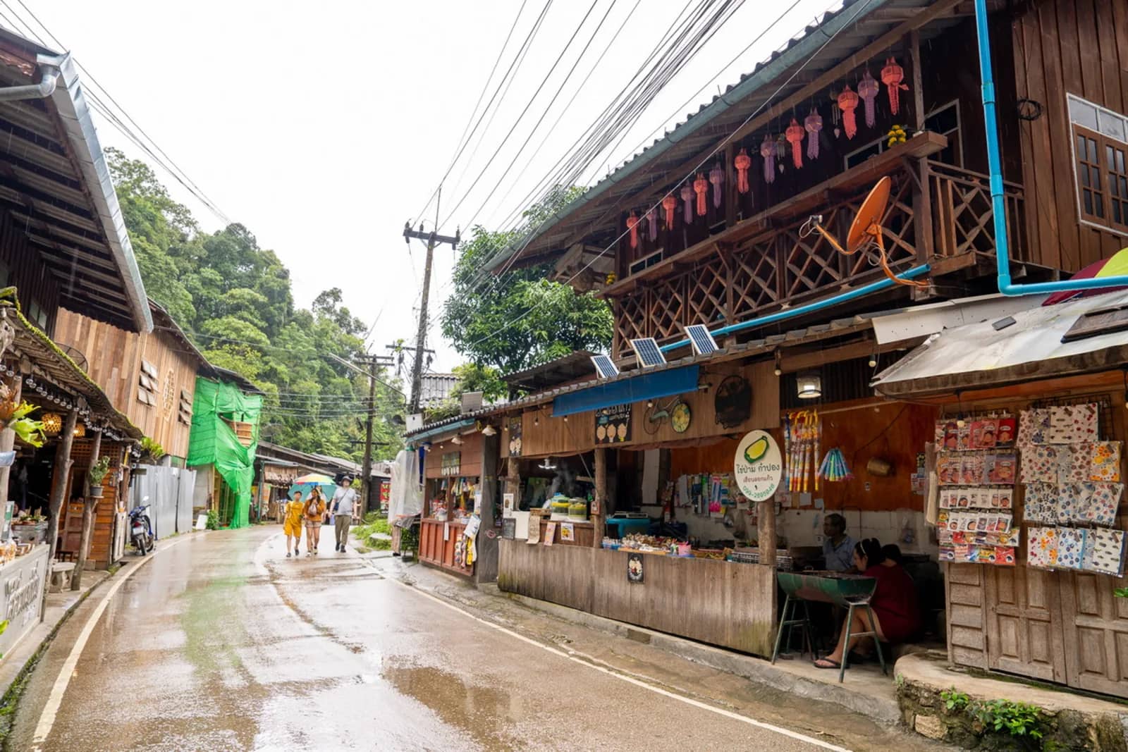 A wet village street lined with wooden shops and hanging lanterns in Mae Kampong