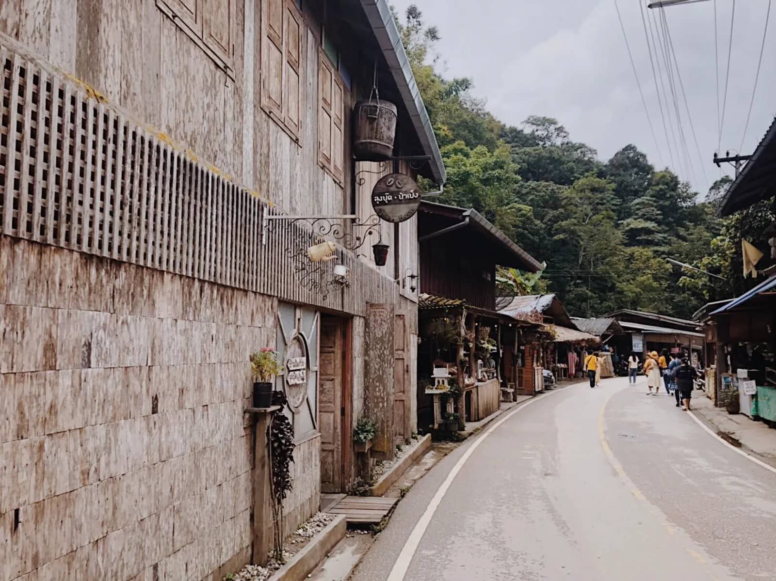 A quiet road winds past weathered wooden buildings and forested hills in Mae Kampong