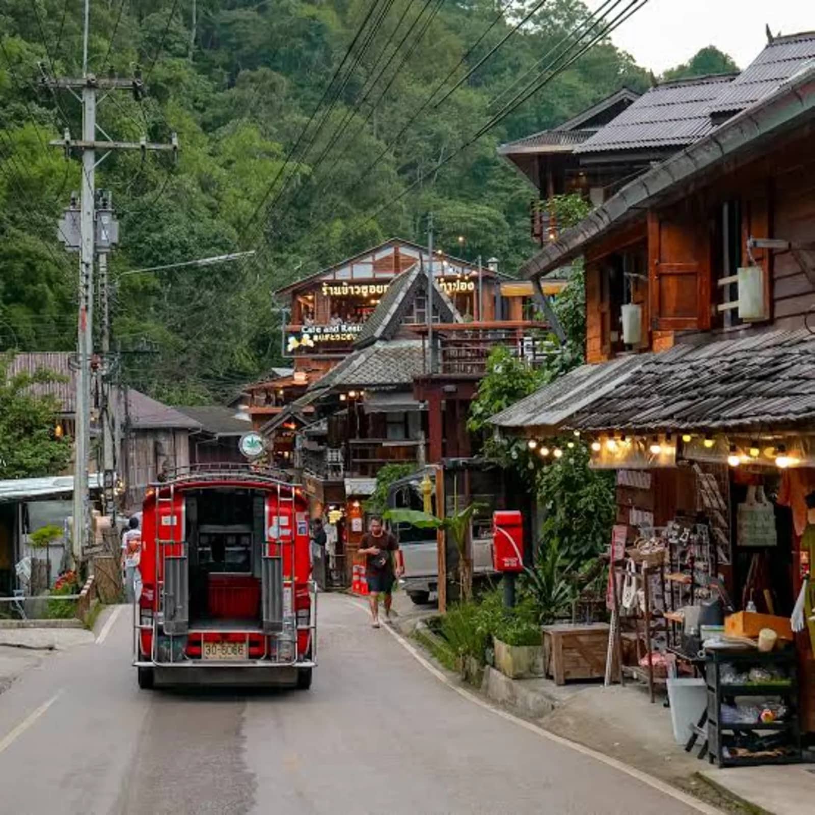 A red songthaew drives up a steep village street lined with wooden shops and houses