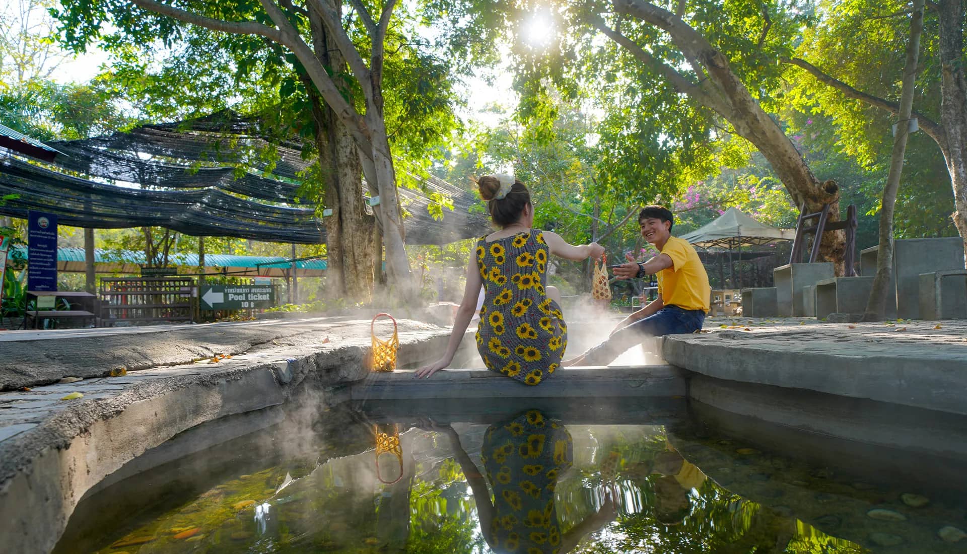 Two visitors sit on the edge of a steaming hot spring pool under shady trees
