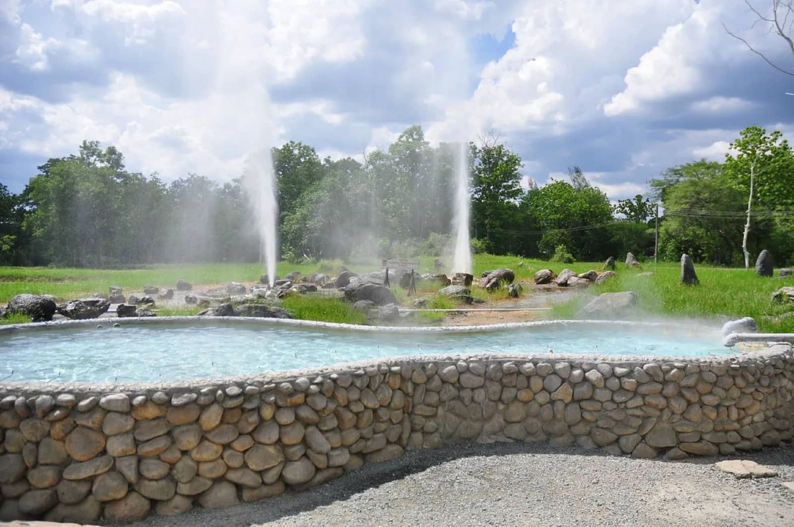 Two geysers shoot steaming water into the air behind a stone-walled hot spring pool