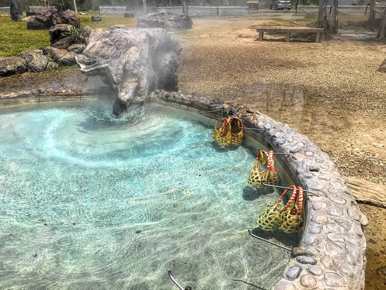 Baskets of eggs hang in a steaming turquoise hot spring pool with a stone rim