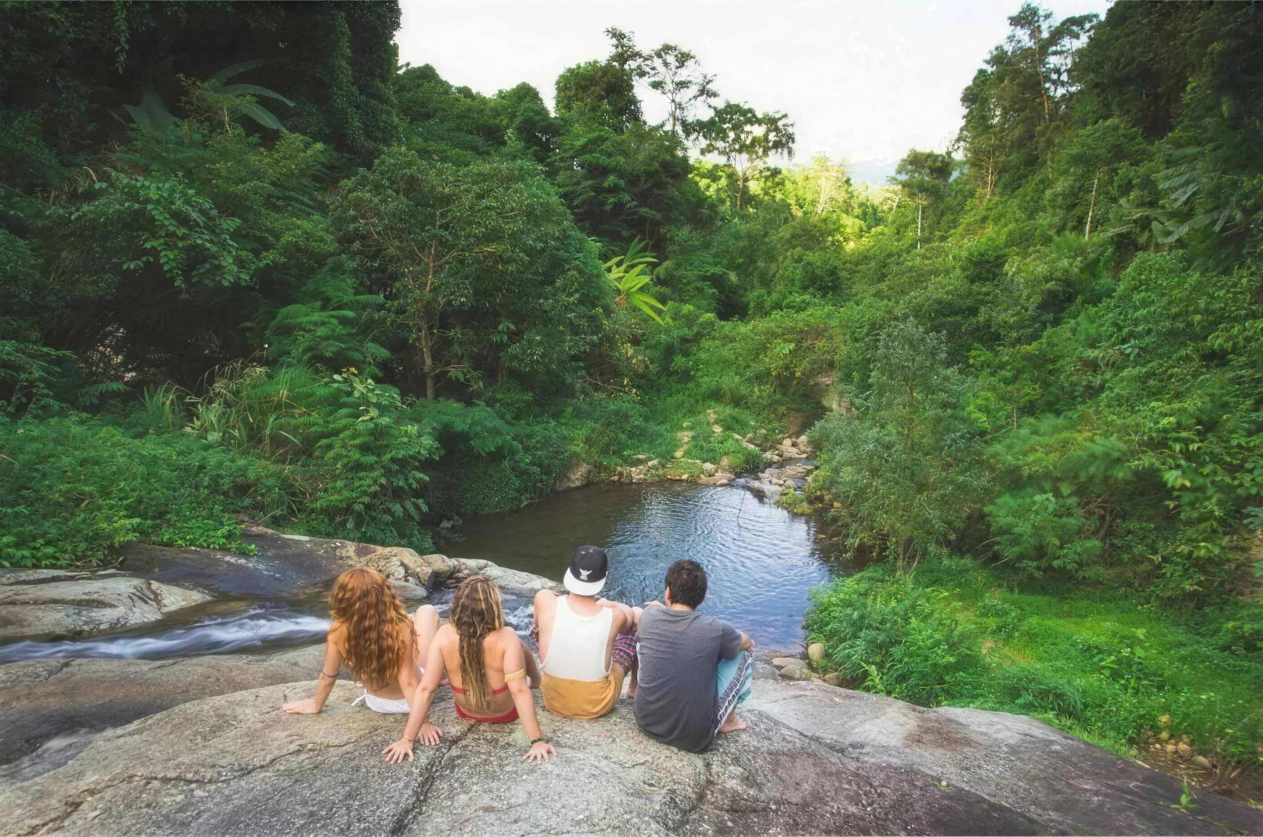 Four trekkers sit on a rock ledge overlooking a green pool surrounded by jungle near Pai