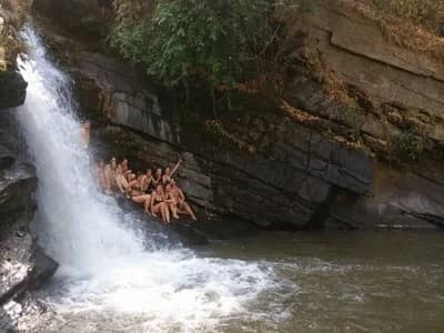 A group of swimmers sits on a rock ledge beside a rushing waterfall on a Mae Wang trek
