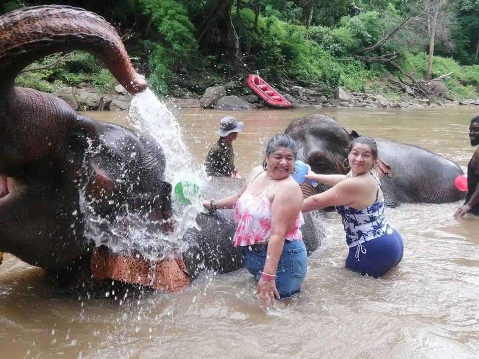 An elephant sprays water from its trunk as two smiling visitors bathe beside it in a river