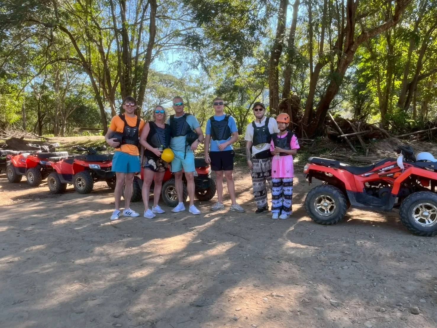 A group of riders in life vests stands between red ATVs in a dirt clearing under shady trees.