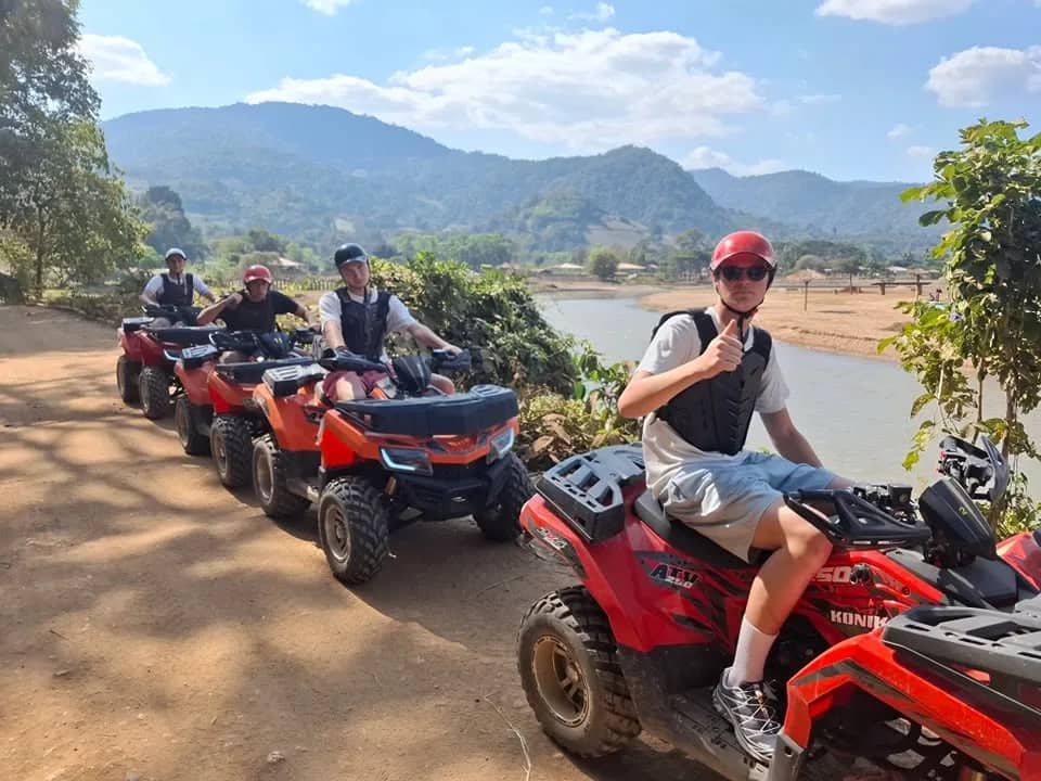A rider gives a thumbs up from a red ATV parked in a line beside a river and mountains
