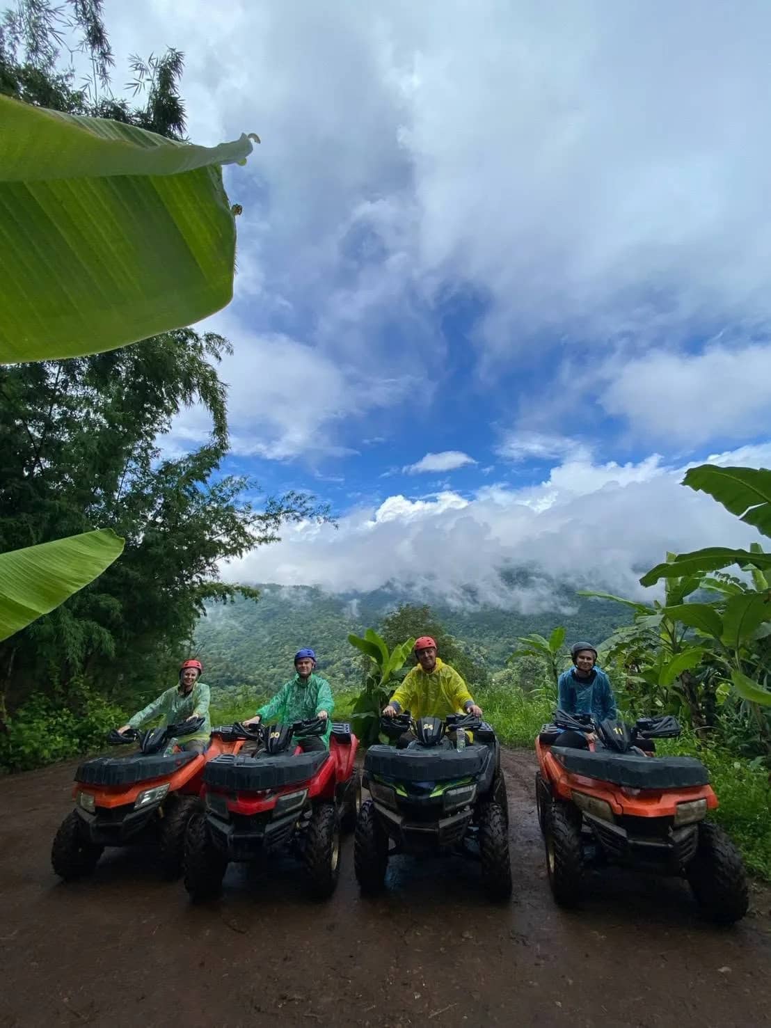 Four riders sit on ATVs lined up on a dirt track with misty mountains behind