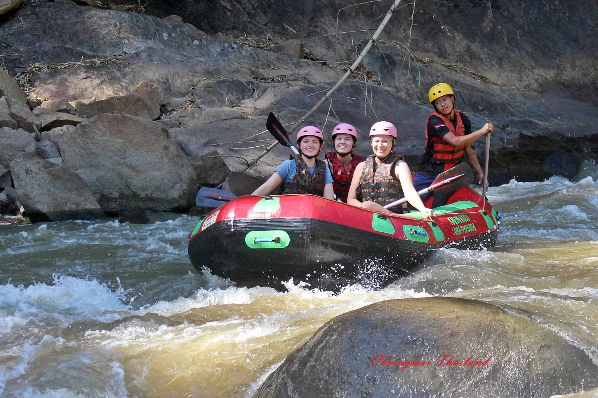 Rafters in helmets paddle a red raft past large rocks in fast-flowing water