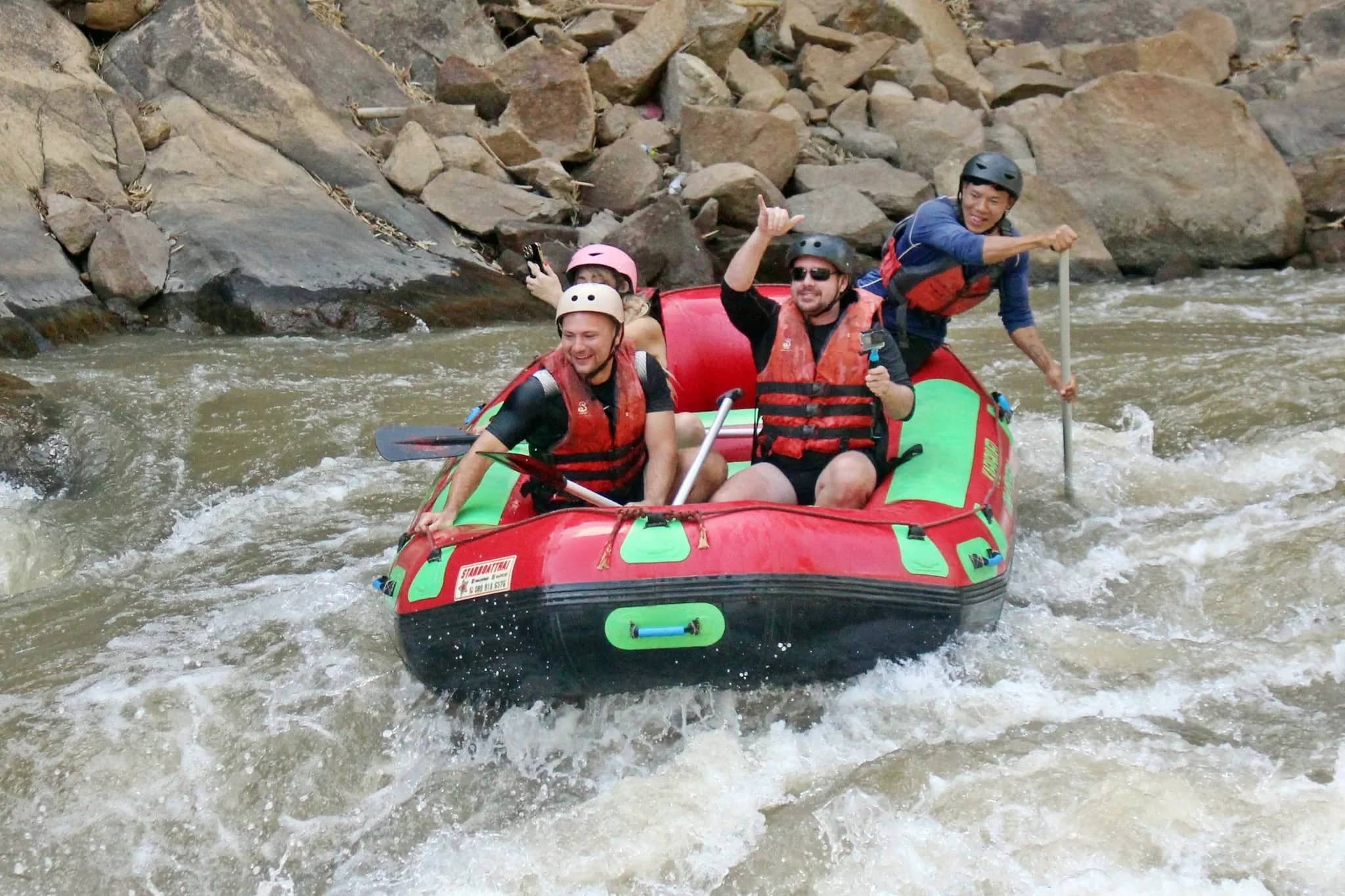 Rafters in helmets cheer as their red raft bounces through rapids below a rocky bank.