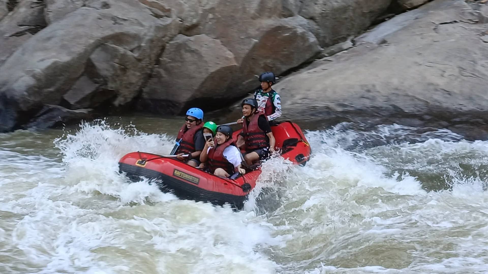 A rafting crew in helmets steers a red raft through foaming whitewater below large rocks