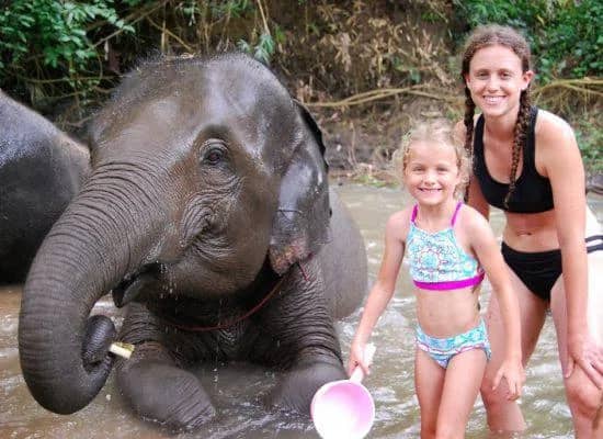 A smiling child and a young woman pose beside a baby elephant lying in a shallow river
