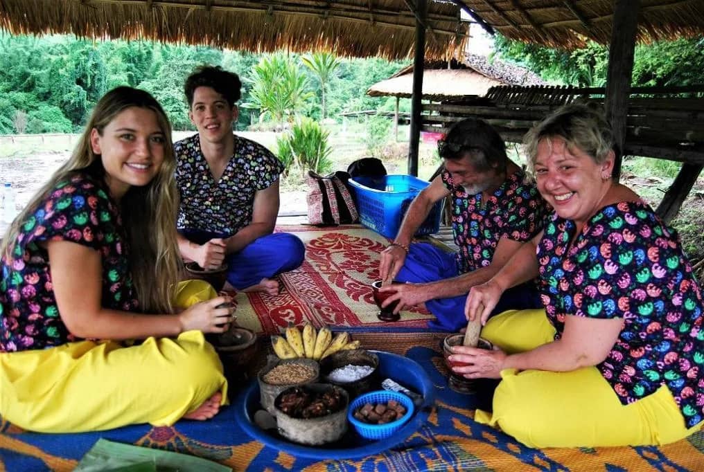 Guests sitting on a mat pound bananas and grains into food balls for the elephants under a thatched roof