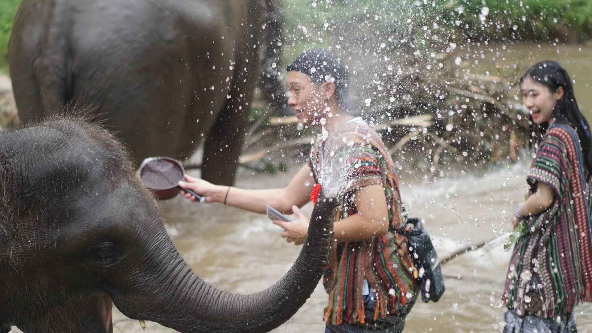 Two visitors in woven tops laugh as a baby elephant sprays water at them in the river.