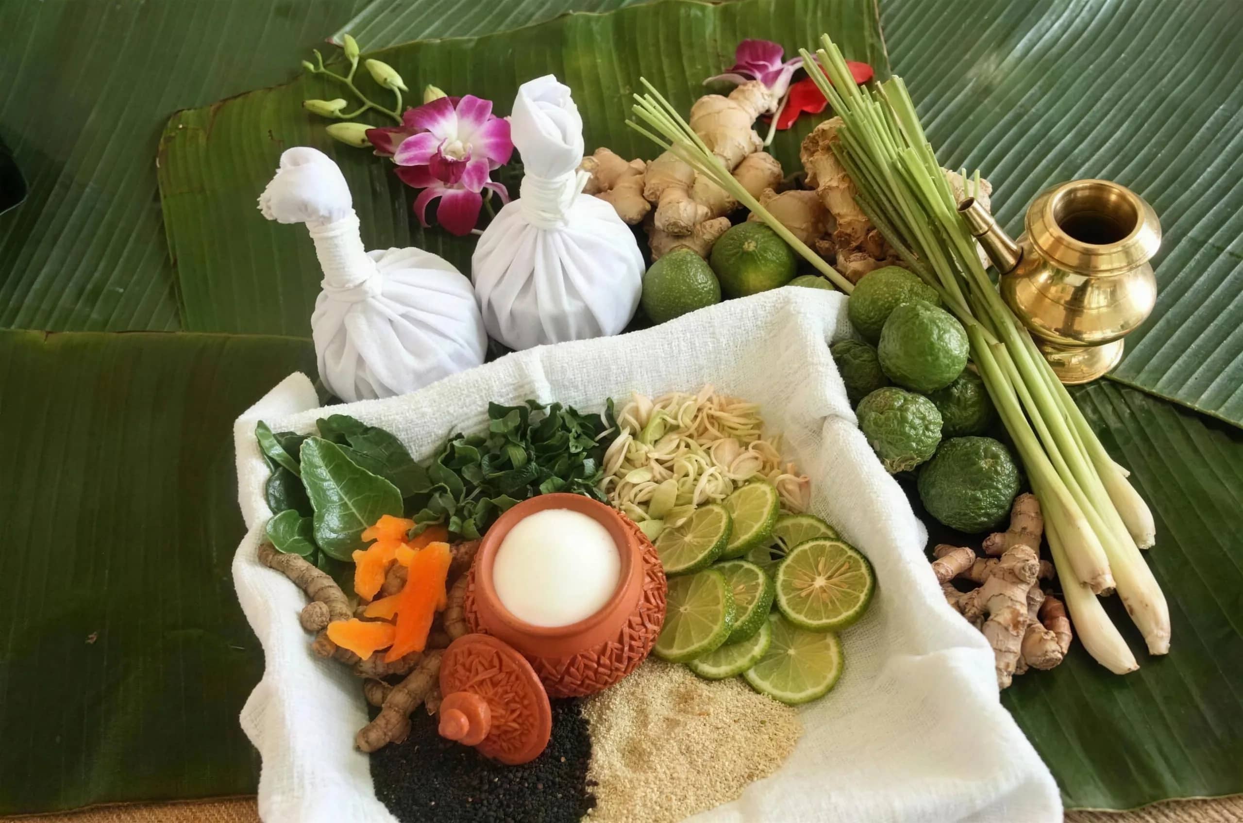 Herbal compress balls, lemongrass, kaffir limes, turmeric and orchids laid out on a banana leaf.
