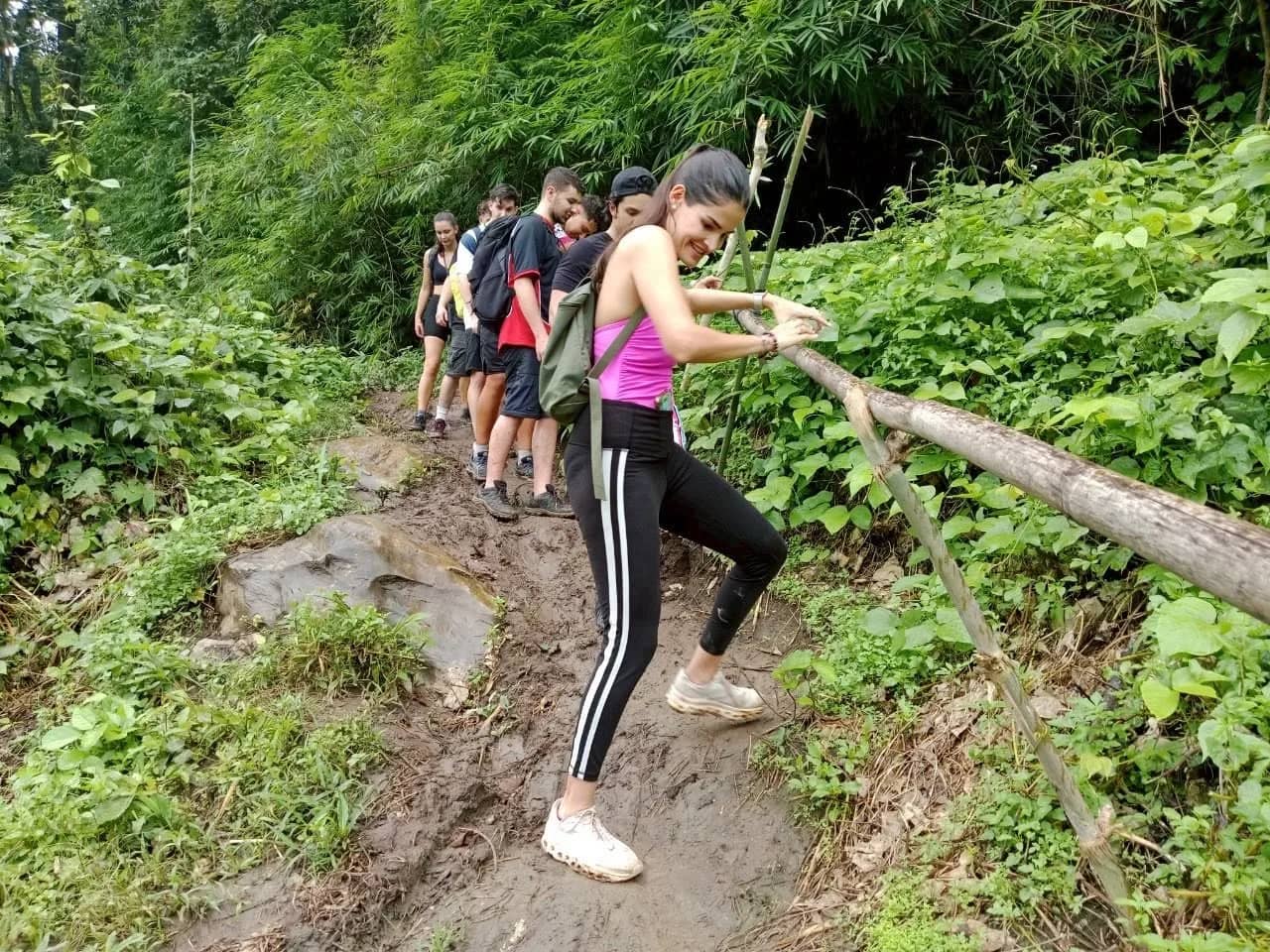Hikers climb a muddy jungle trail, one holding a bamboo railing beside the path near Chiang Mai
