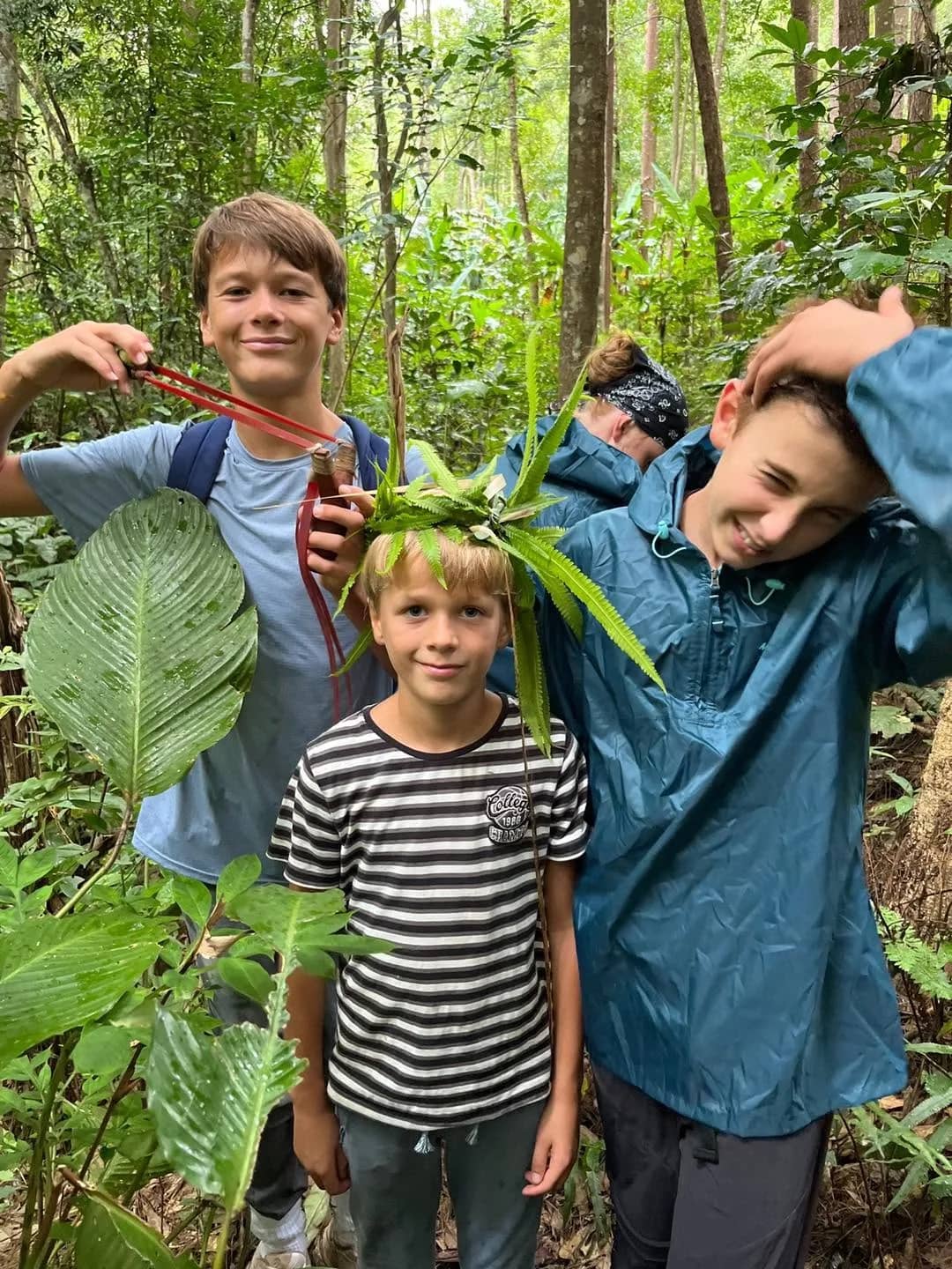Three boys play in the jungle, one wearing a crown of green leaves on his head.