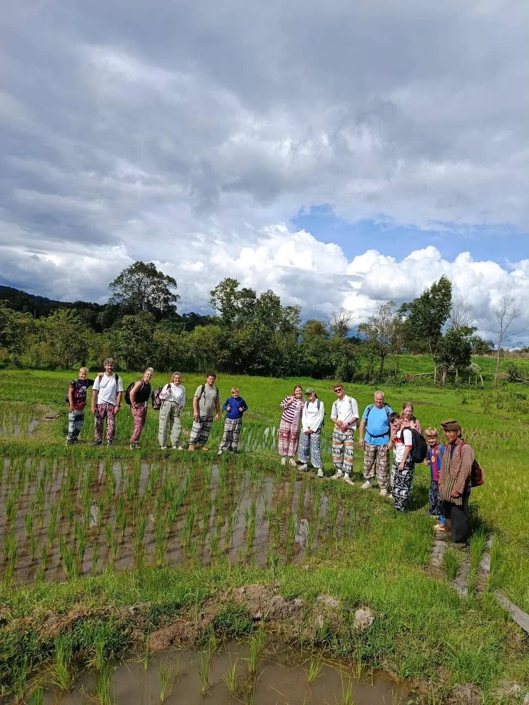 A long line of trekkers walks along the edge of a flooded rice paddy under a cloudy sky.