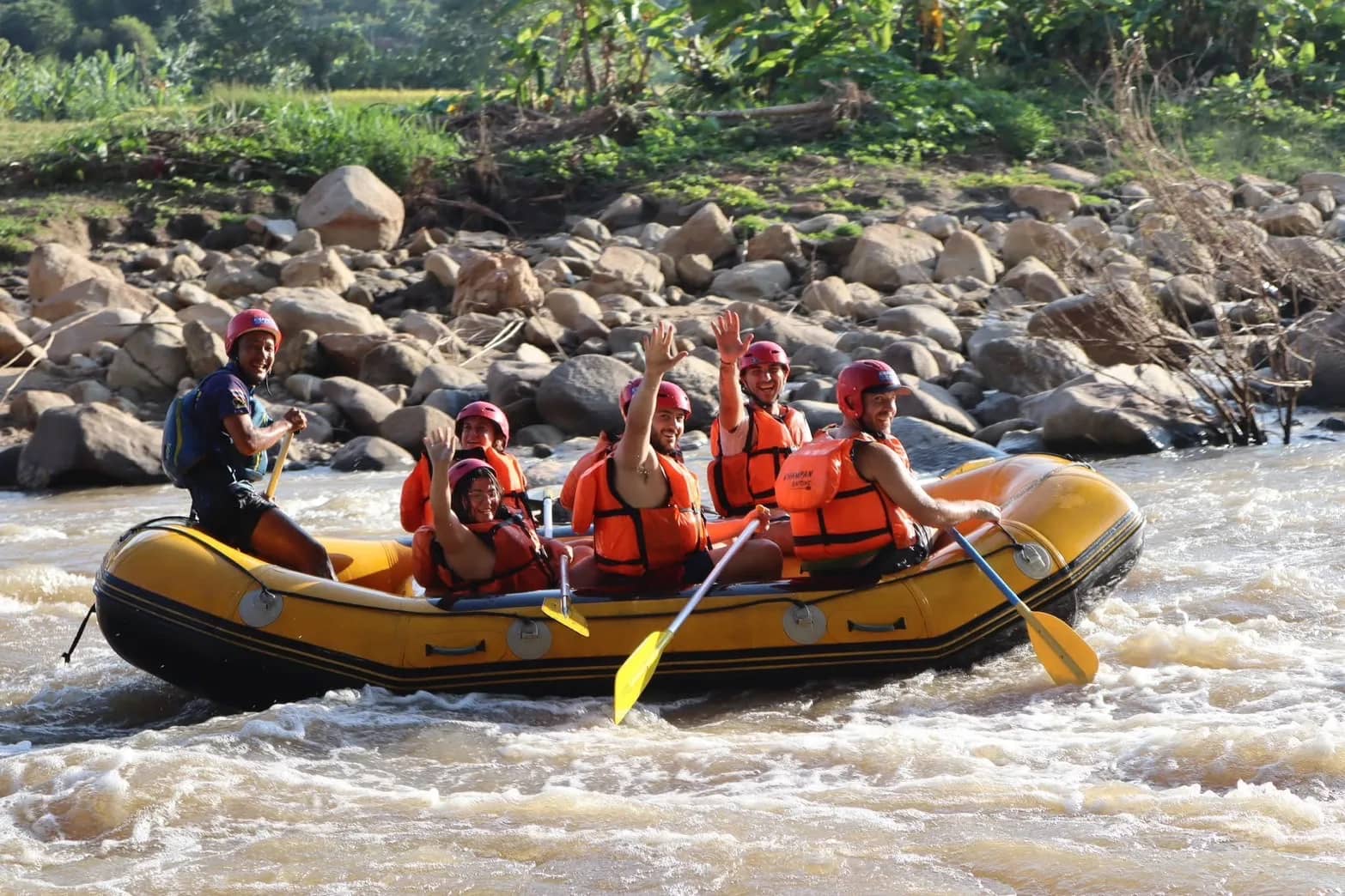 Rafters in red helmets and orange life jackets wave from a yellow raft on a rocky river