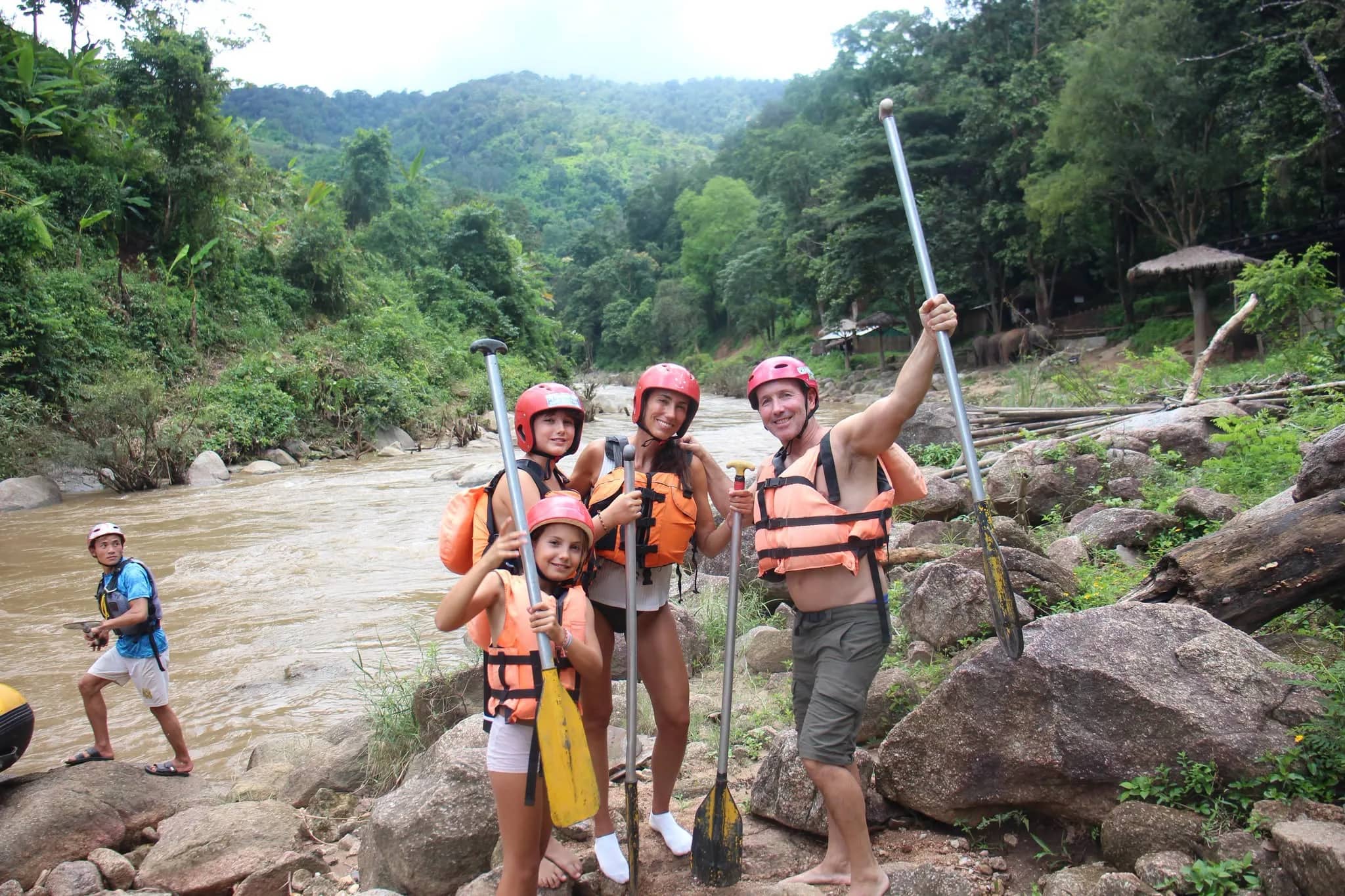 A group in helmets and life jackets raise their paddles on the rocky bank of a jungle river
