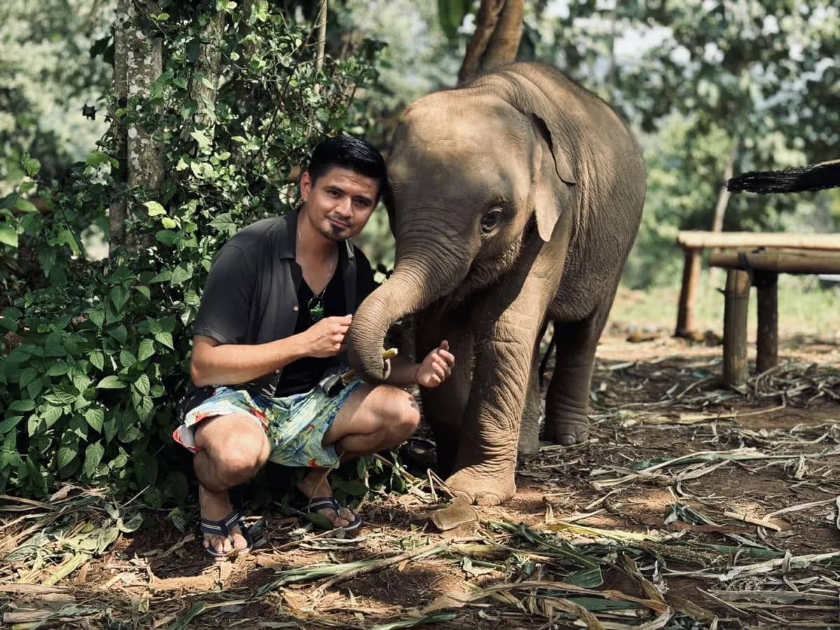 A visitor crouches beside a baby elephant that rests its trunk on his arm in the forest