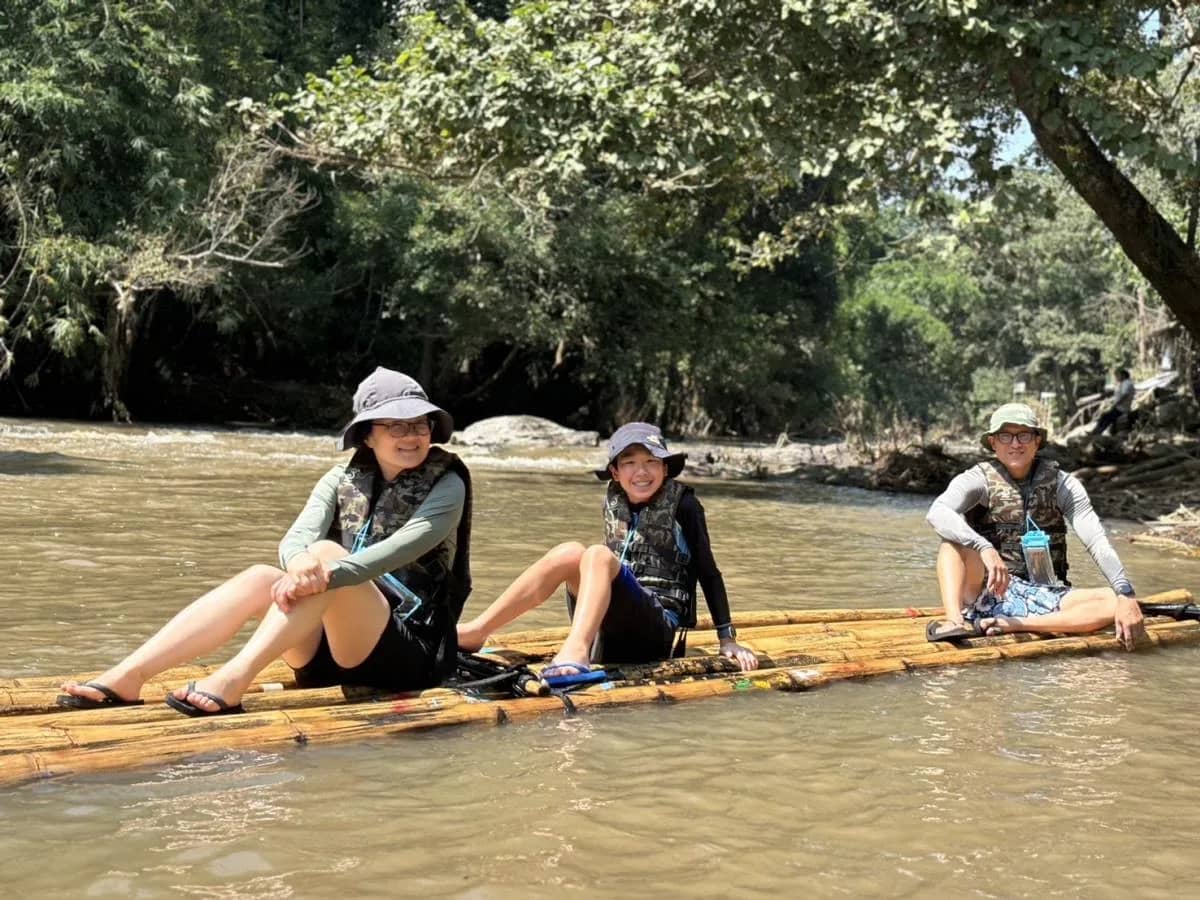 Three guests in sun hats and life jackets sit on a bamboo raft drifting down a river