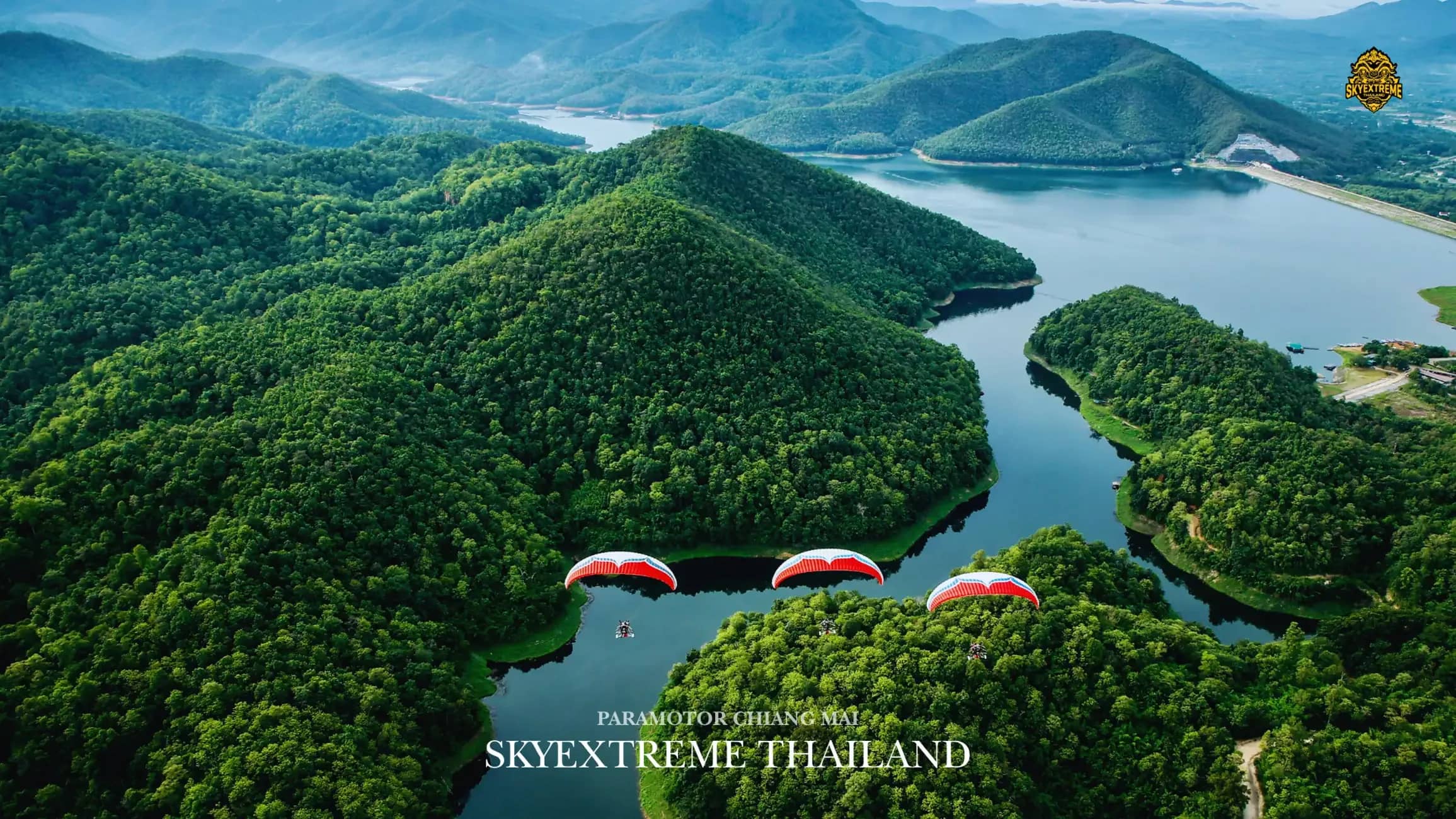 Three paramotor wings fly low over a winding reservoir between green forested hills