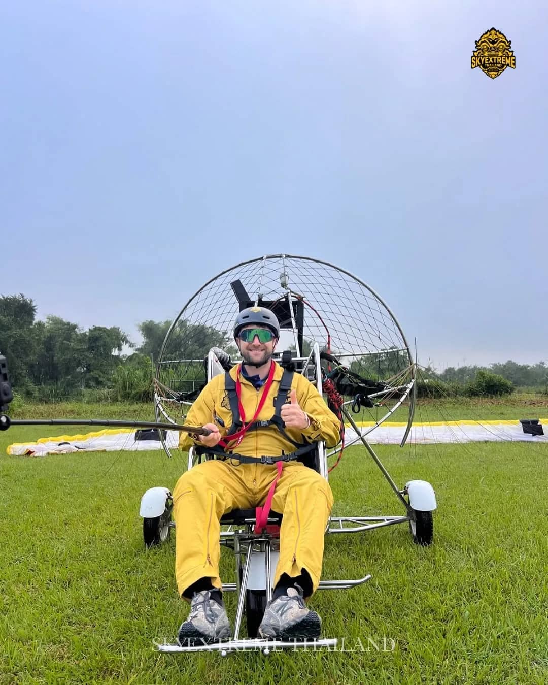A smiling pilot in a yellow flight suit gives a thumbs up seated in a paramotor trike on grass