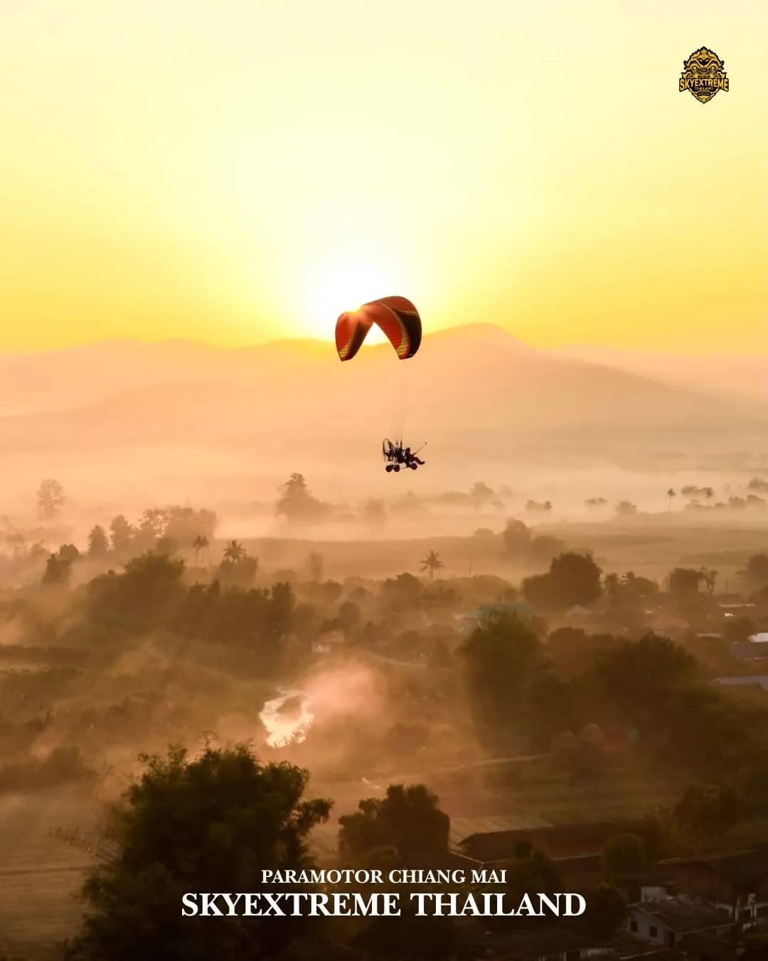A paramotor flies through a golden sunrise above a misty valley of trees and villages