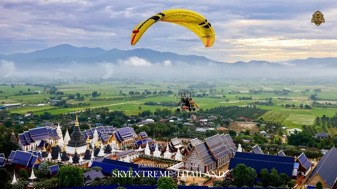 A yellow paramotor wing flies above a large temple complex and green fields below misty mountains