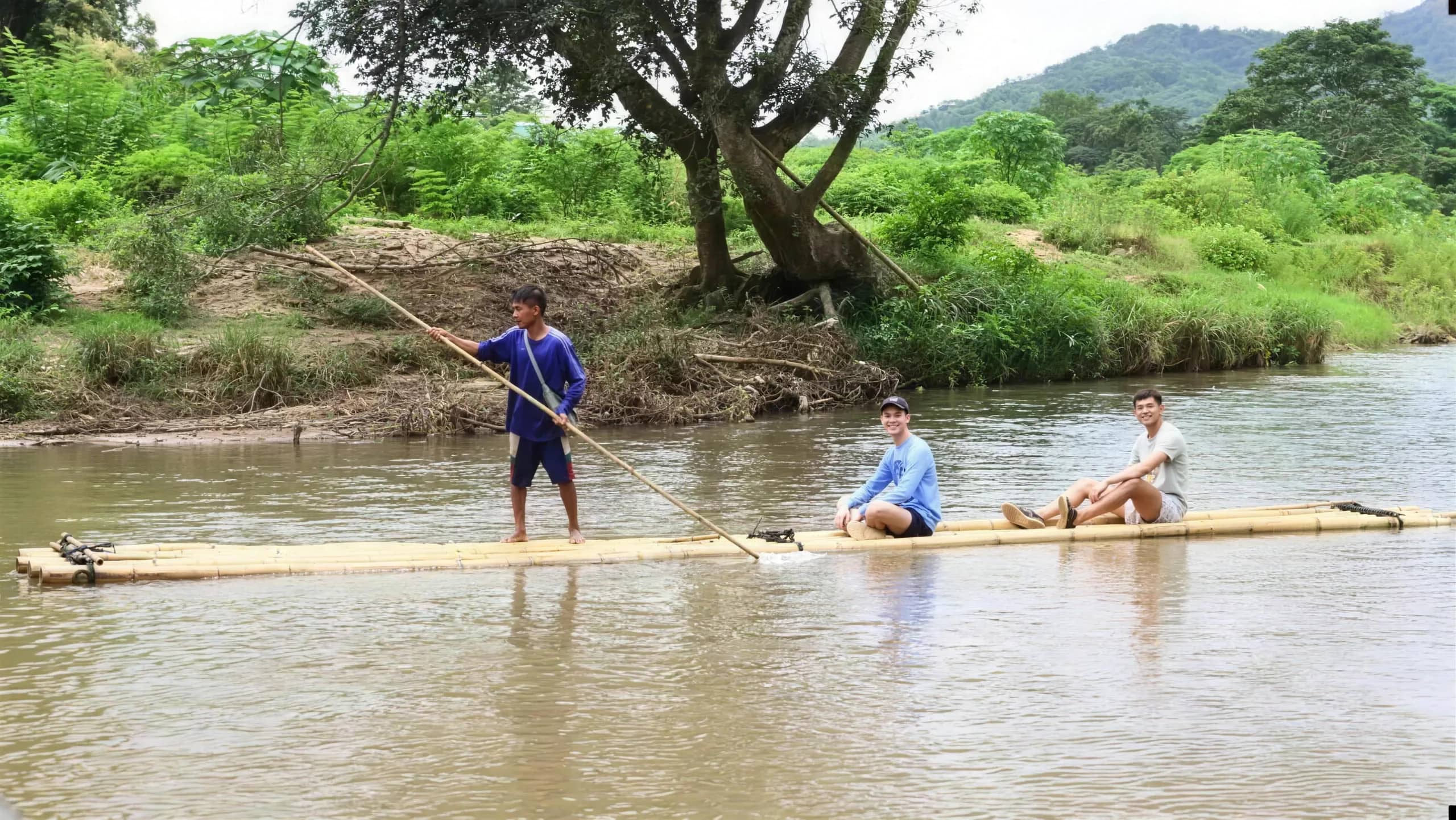 A guide poles a long bamboo raft carrying two visitors down a wide river with hills behind