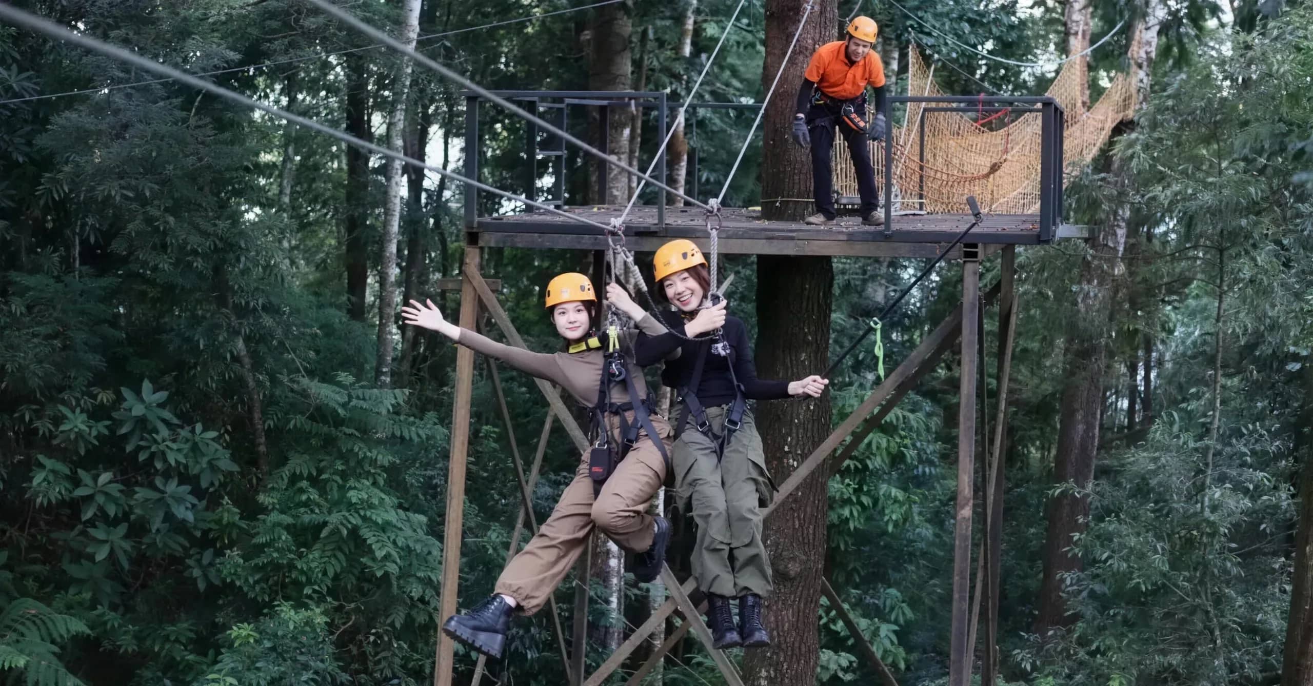 Two riders in helmets wave as they leave a tree platform on a zipline at Skyline Jungle Luge