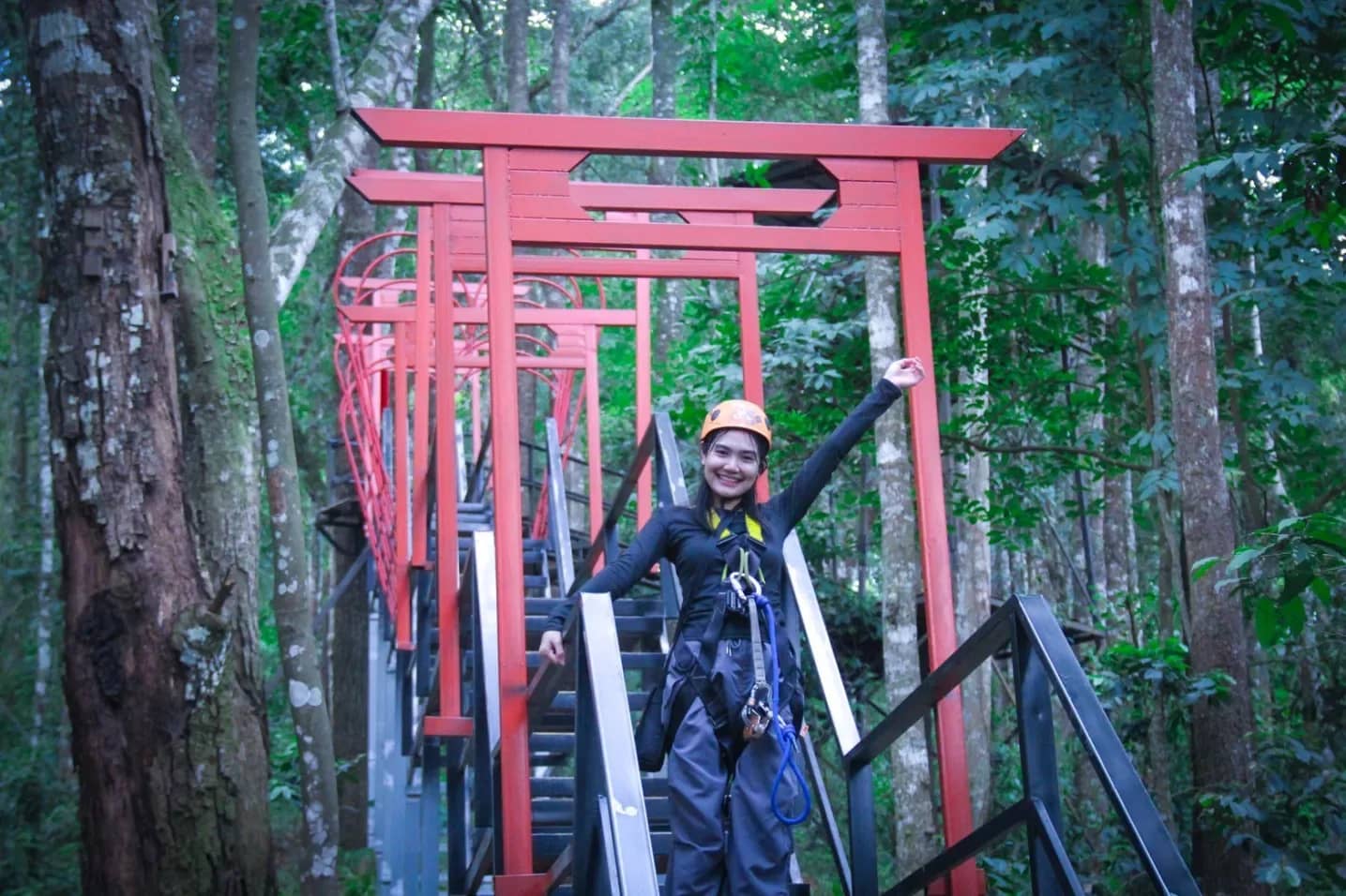 A rider in climbing gear waves from a steel stairway under red torii gates in the forest