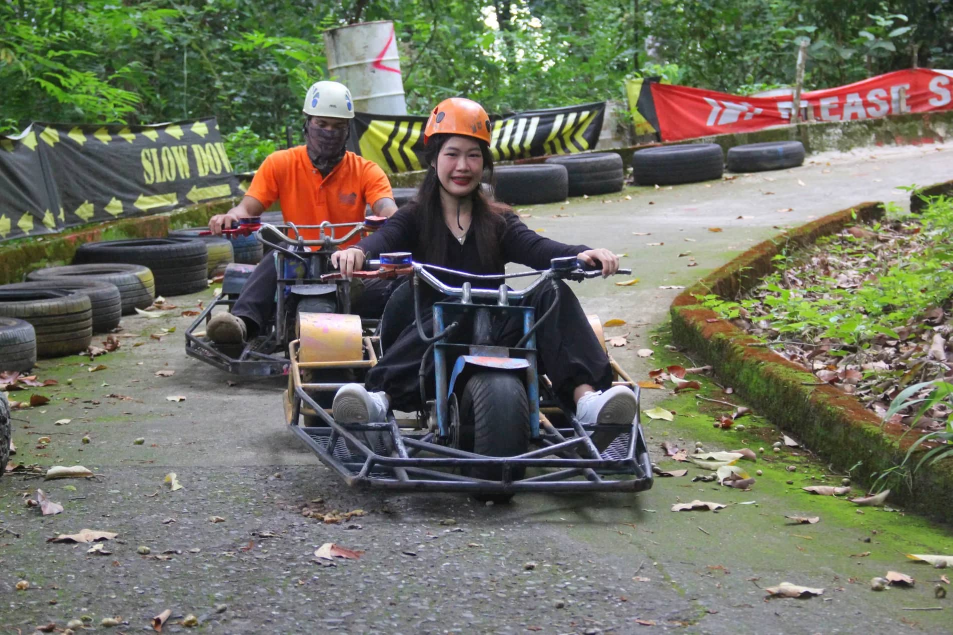 Riders in helmets steer small luge karts down a curving track lined with tyres in the forest