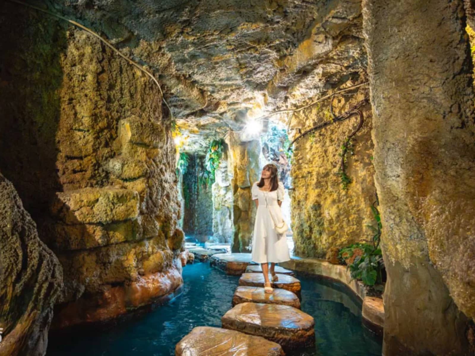 A visitor in a white dress walks across stepping stones over turquoise water inside a rock cave.