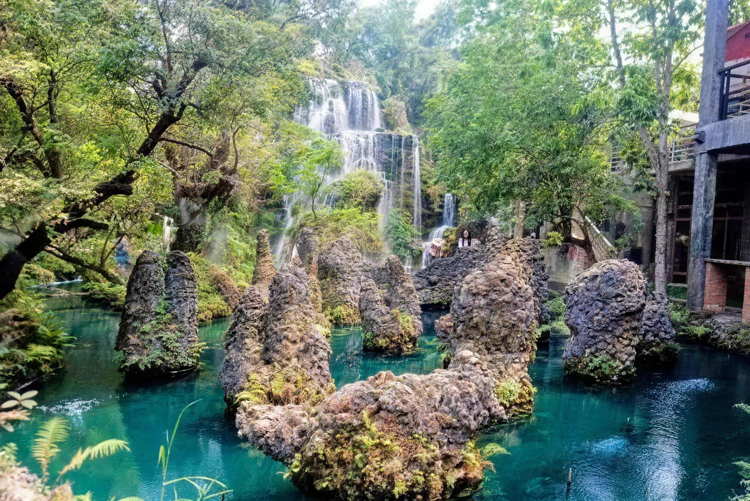 Jagged rock formations stand in a blue-green pond in a landscaped garden beside wooden buildings.