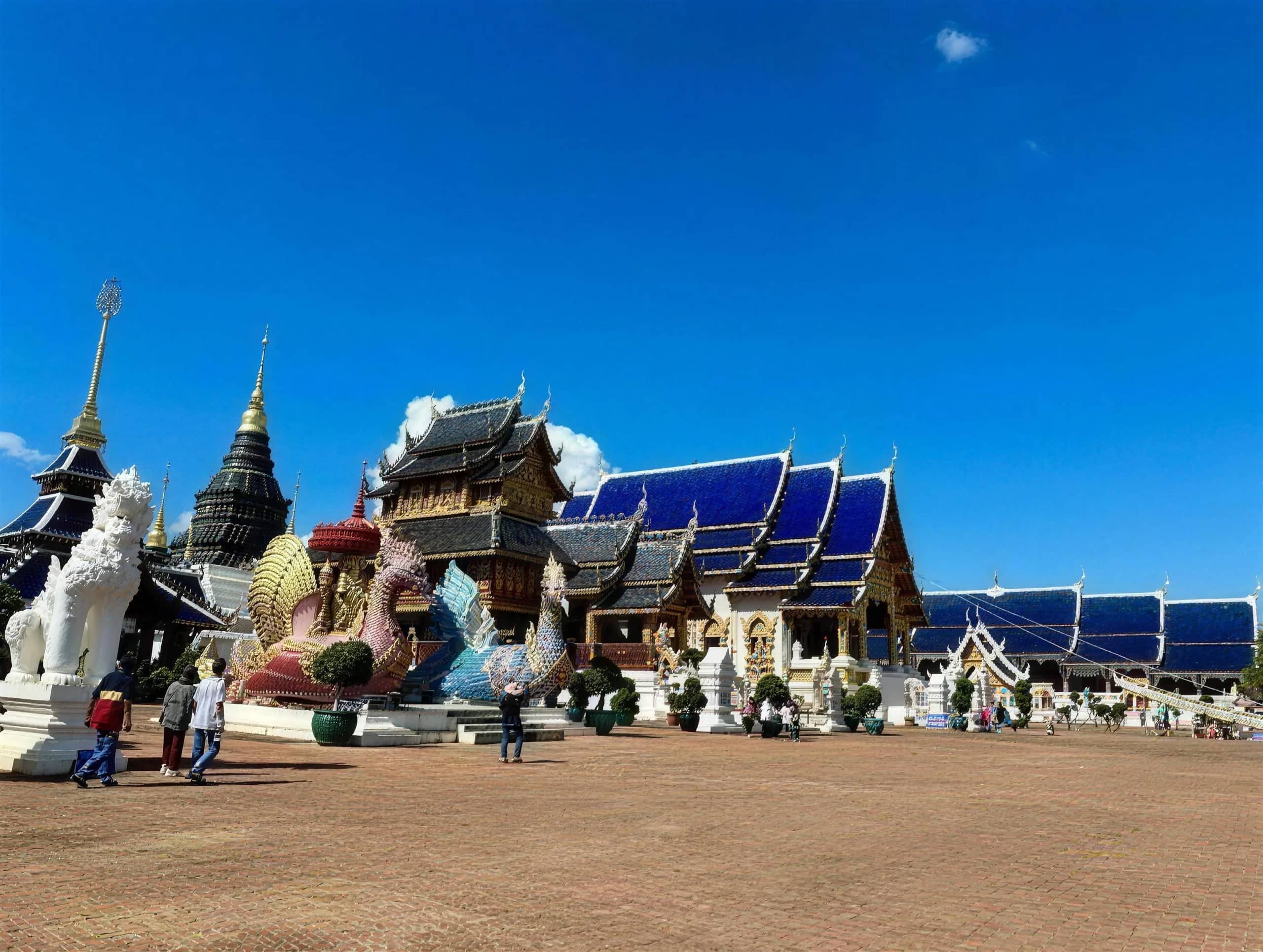 Visitors walk across the open courtyard of Wat Ban Den with guardian statues and blue-roofed halls