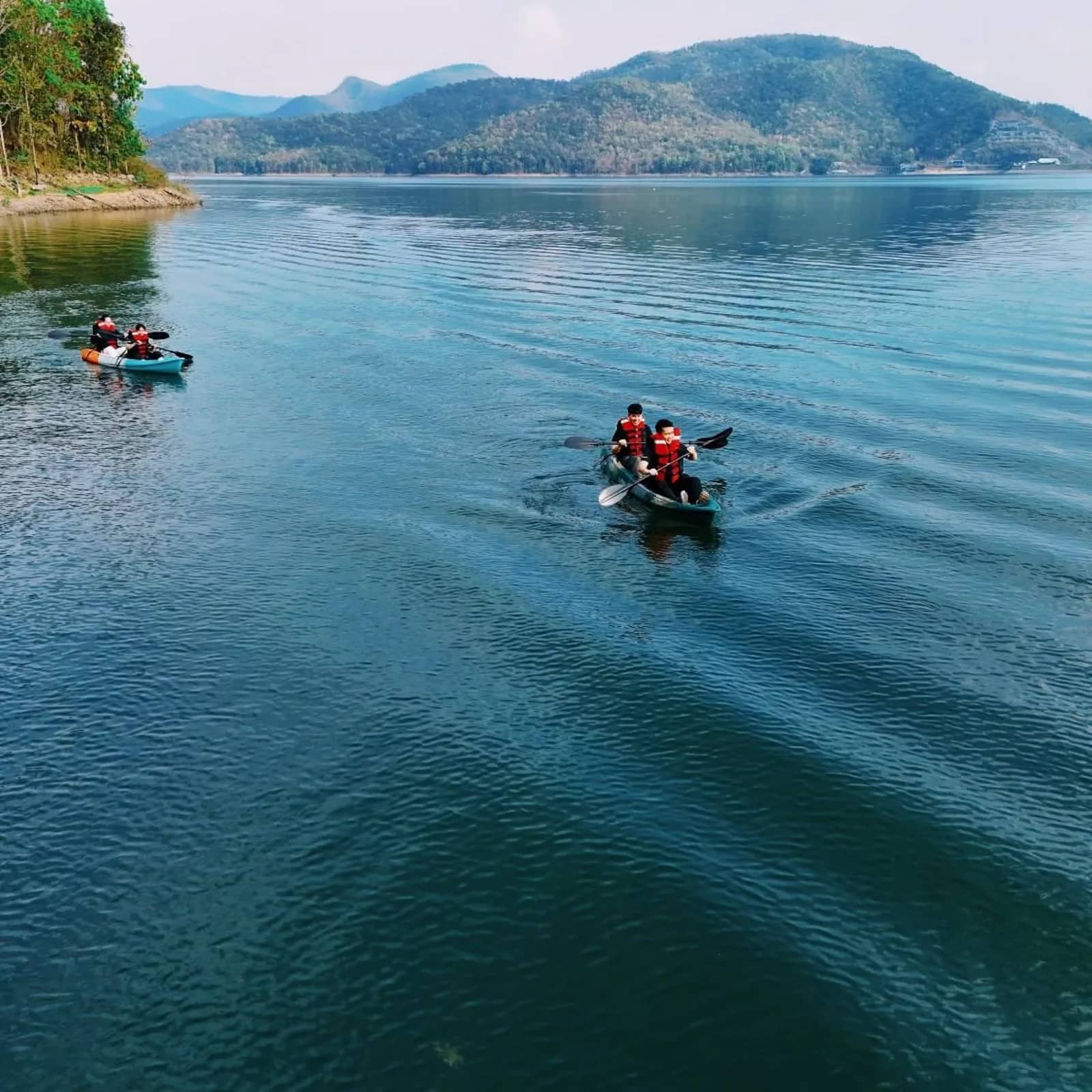 Two kayaks with paddlers in life jackets glide across a calm blue lake below forested hills