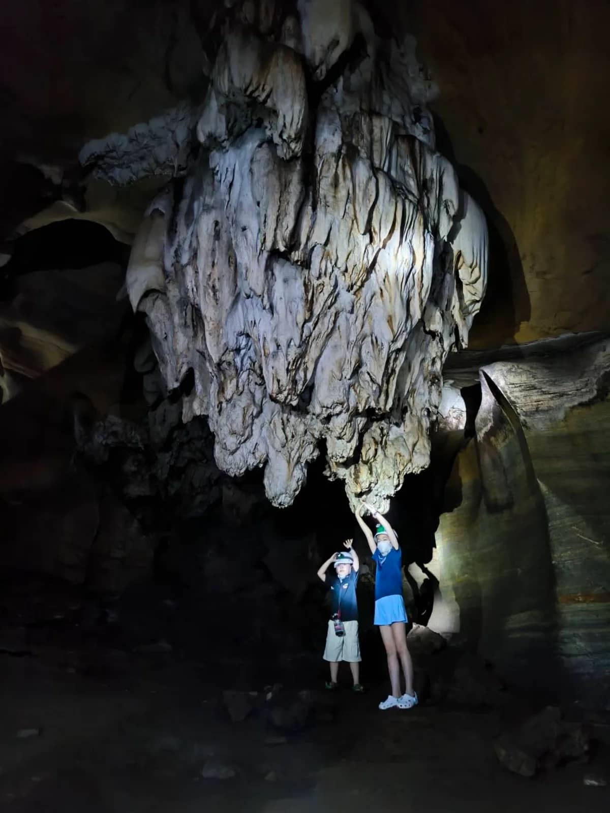 Two visitors look up at huge white rock formations hanging from the ceiling of a dark cave
