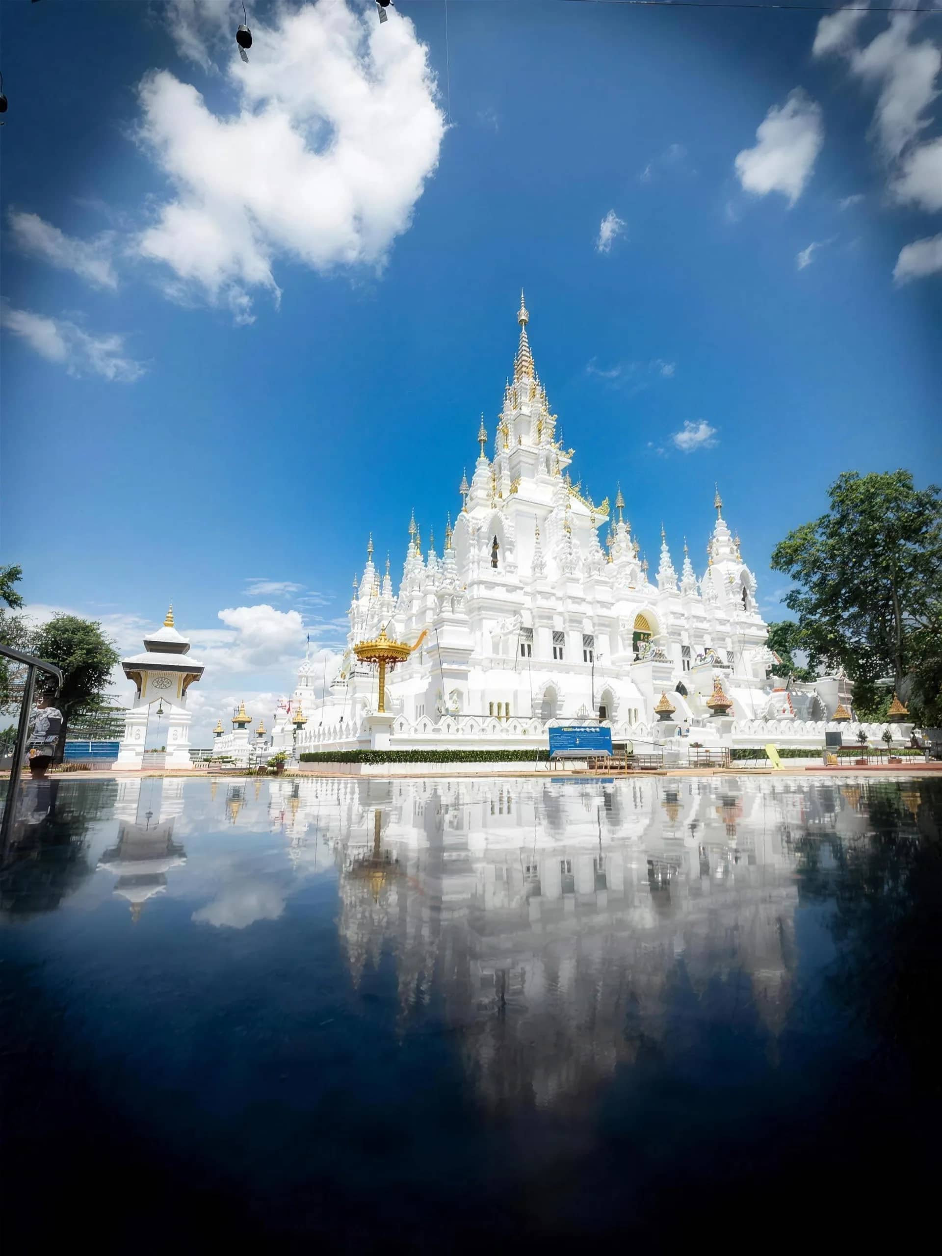 A white temple with tall spires is mirrored in a still pool under a blue sky with clouds