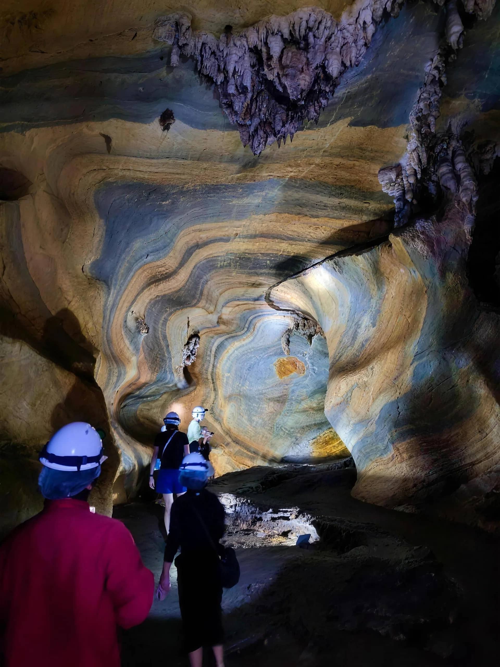 Visitors in helmets walk through a cave with swirling bands of colourful rock
