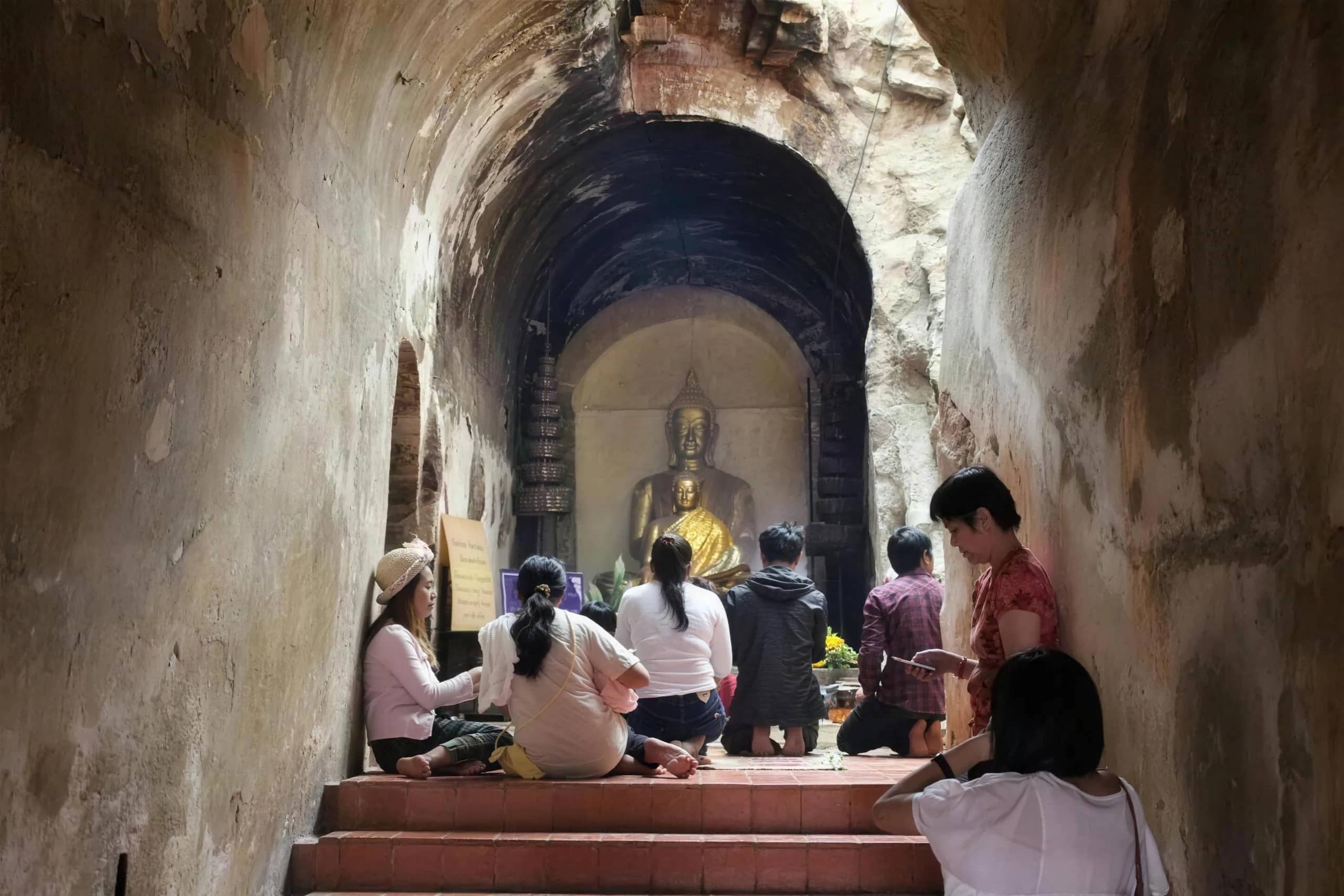 People kneel in prayer before a golden Buddha at the end of an ancient brick tunnel at Wat Umong