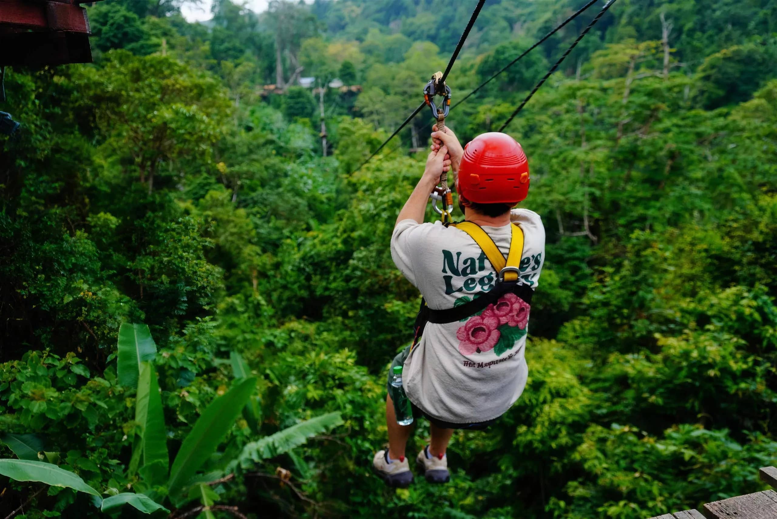 A rider in a red helmet holds the harness while gliding away over the dense jungle.