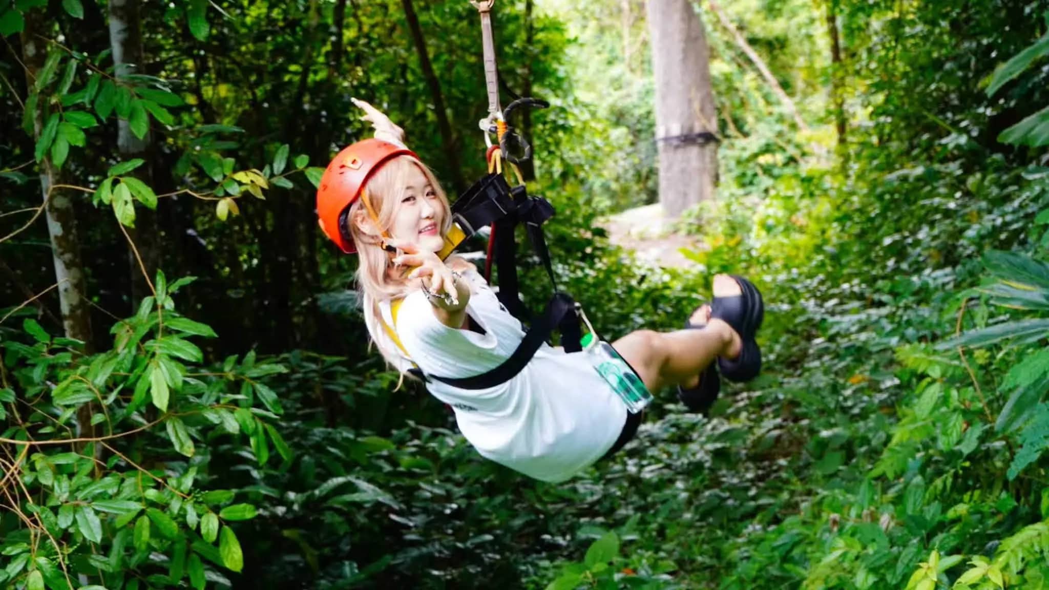 A rider in an orange helmet smiles and points while hanging from a zipline among jungle trees.