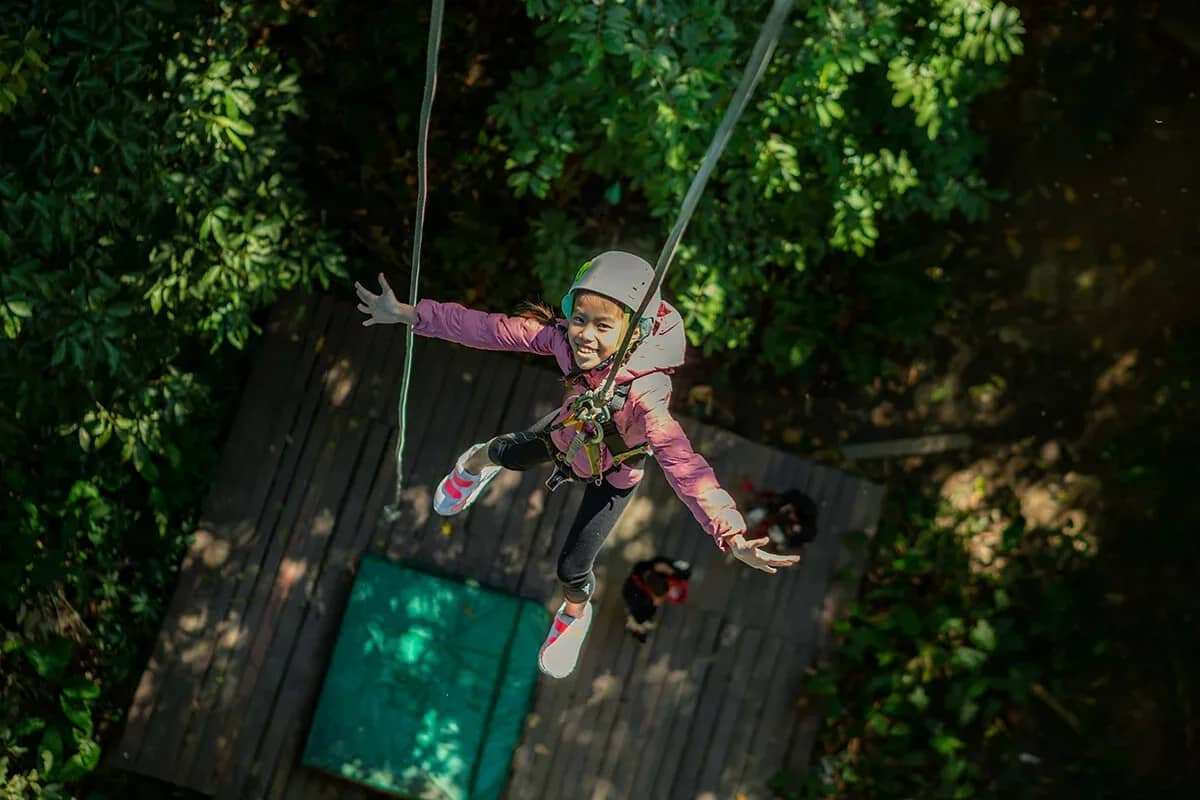 A smiling child in a helmet and harness spreads her arms on a zipline above a wooden platform.